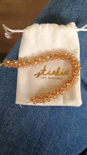 Recensent toont de gouden Stiekie kralen haarelastiek die op het witte merkzakje ligt.
