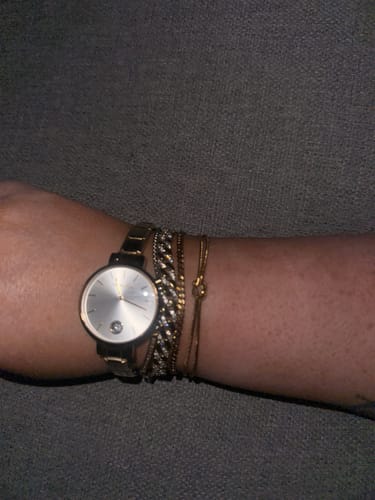 Reviewer's wrist showing a gold knot bracelet worn with a watch and other stacked bracelets.