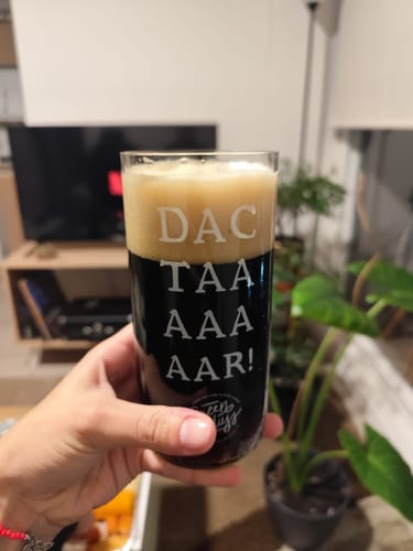 Reseñador sosteniendo el Vaso Dactar con una bebida oscura y espuma, mostrando el texto 'DAC TAA AAA AAR!'.