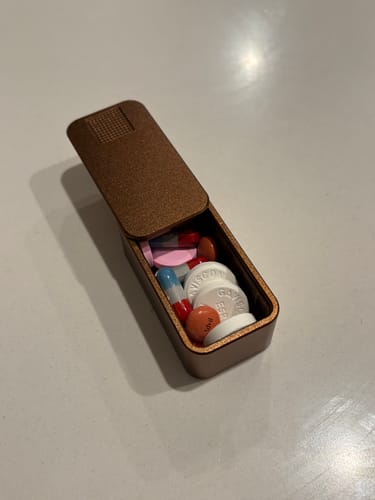 Customer's coffee-colored metal pill case, open to show a variety of pills and capsules inside.