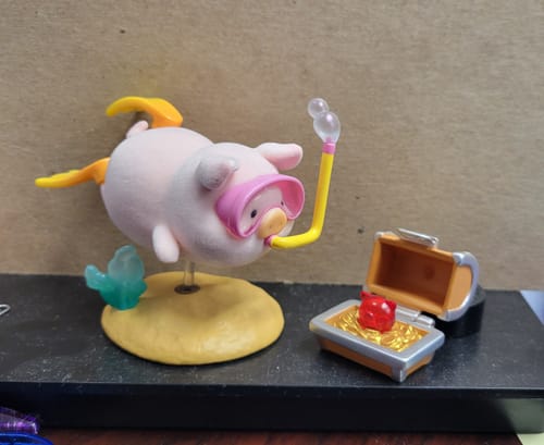 Reviewer's Lulu The Piggy figure in pink snorkel gear, swimming towards a treasure chest with a red pig-shaped gem.