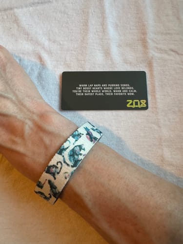 A reviewer wears the Cat Mom Bracelet on their wrist, showing its muted cat pattern next to the accompanying card.