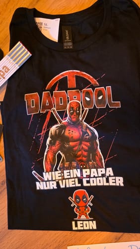 Kunde zeigt das personalisierte schwarze Dadpool-T-Shirt, das auf einer Holzoberfläche ausgebreitet ist.