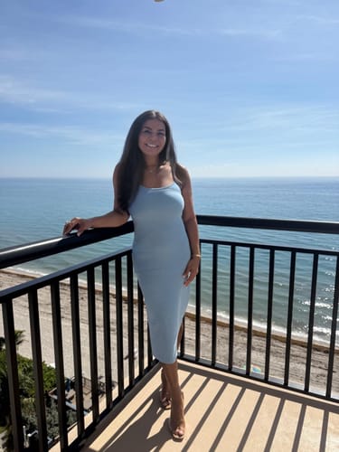Reviewer standing on a sunny balcony, wearing the Victoria Clear Strap Mule Heels with a blue dress.