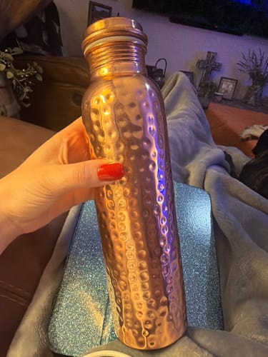 Reviewer's hand holding the shiny, hand-hammered Pure Copper Water Bottle in a living room setting.