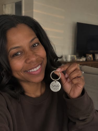 Reviewer smiling while holding up the silver, circular "Smile | It Looks Good On You Keychain" in their home.