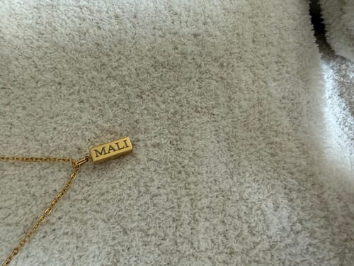 Customer's gold Discrete Mini Urn Bar Pendant engraved with 'MALI', seen close-up on a soft surface.