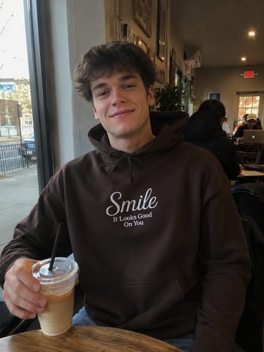 A smiling reviewer in a coffee shop wearing the brown 'Wear Your Smile Hoodie'.