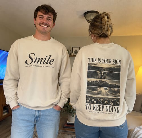 Reviewers wearing the cream-colored Smile Crewneck, showing the 'Smile' text on the front and the graphic design on the back.