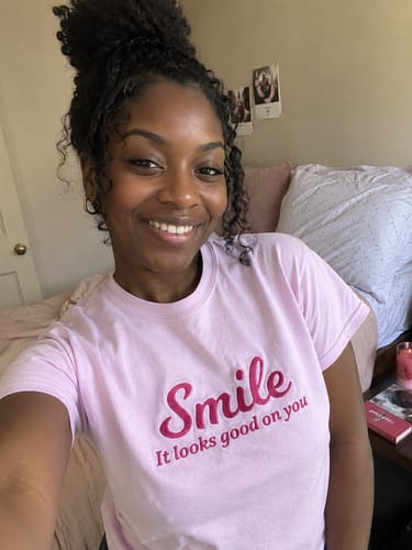 Customer smiling while wearing the light pink Wear Your Smile Shirt in a bedroom.