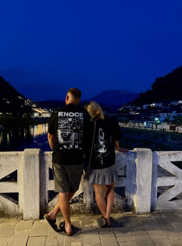Customer wearing the black EMBRACE THE STRUGGLE OVERSIZED HEAVY TEE on a bridge at night, viewed from behind.
