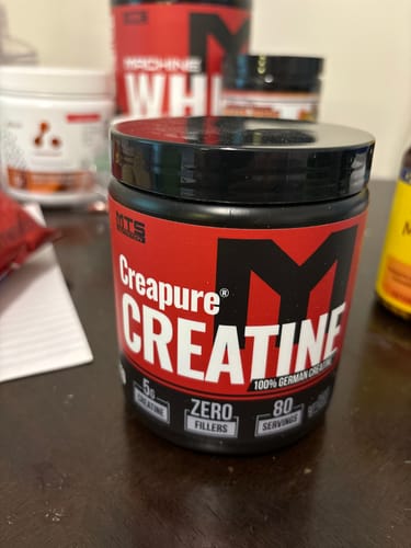 Customer's black tub of MTS Creapure Creatine powder shown close-up on a desk with other supplements in the background.