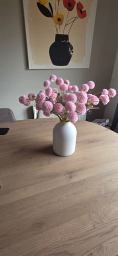 Recensent toont de roze Zijden Kiku Bloemen in een witte vaas op een houten tafel.