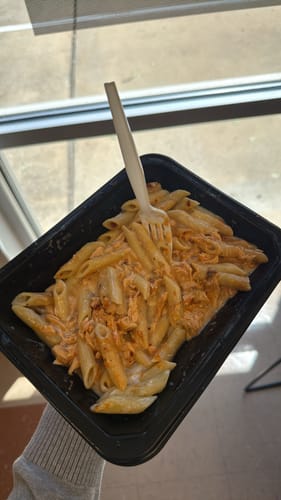 Customer holding a black container of prepared Salmon Alfredo pasta with a fork.