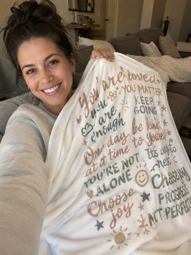 Reviewer smiling on a couch while holding up the white Sherpa blanket with its colorful, inspirational text.