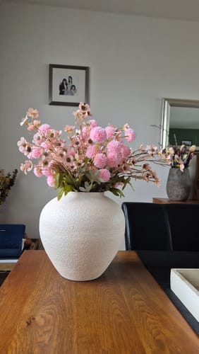 Recensent toont de crèmekleurige Sena vaas op een houten tafel, gevuld met een boeket roze bloemen.