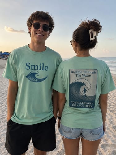 Two customers on a beach wearing the Breathe Through The Waves Shirt, showing the front 'Smile' and back 'Breathe' designs.