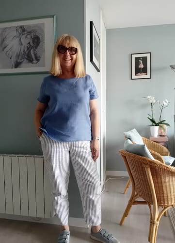 Cliente portant la blouse Emeline en lin bleu méditerranéen avec un pantalon rayé dans son salon.