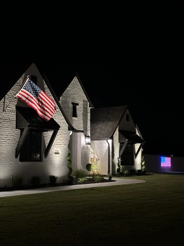 Reviewer's Prestige House Flag Pole mounted on their illuminated brick house at night, displaying a large American flag.