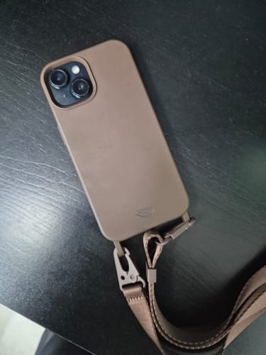 Avaliador mostrando a Alça Aurora Marrom Café conectada a uma capa de celular em uma mesa escura.