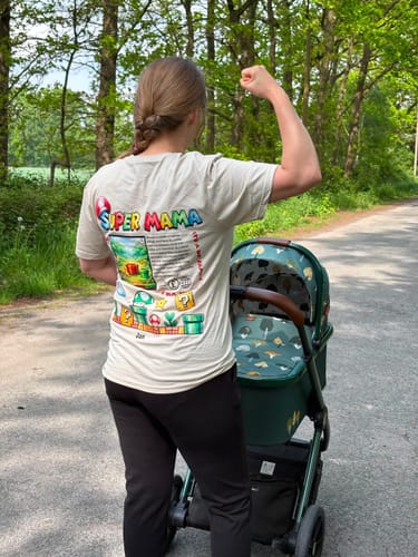 Kundin, die FastLane+ bewertet, posiert im Freien neben einem Kinderwagen und spannt einen Muskel.