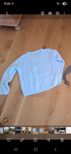 Rezensent zeigt den hellblauen, flauschigen Cardigan SUMMER WARM UP 2.0, der auf einem Holzboden liegt.