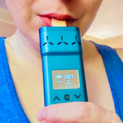 Reviewer holding the blue X ROYAL EDITION vaporizer to their lips. The screen shows it is on and set to 390°F.