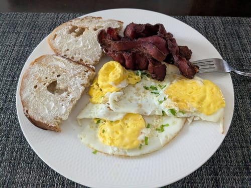 Reviewer's cooked beef bacon served on a white plate with fried eggs and buttered toast.