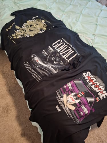 Customer displays three black graphic t-shirts from their MYSTERY CASH BOX laid out on a bed.