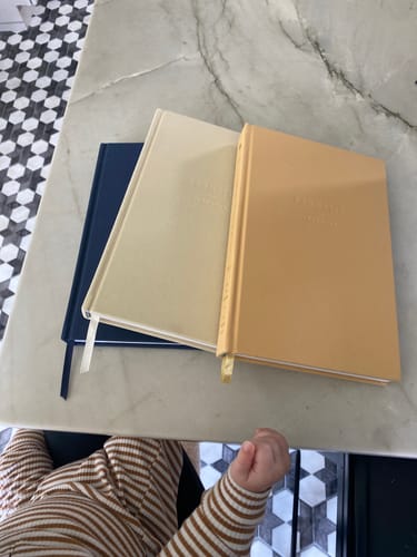 Reviewer's three Childhood History journals in navy, cream, and tan on a countertop with a baby's arms visible in the foreground.