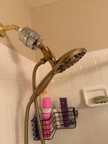 Reviewer's silver Shower Head Filter installed in a tiled shower, connecting the pipe to a bronze shower head.