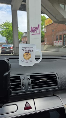 Rezensent zeigt die personalisierte OLDTIMER-Tasse auf dem Armaturenbrett eines Autos.