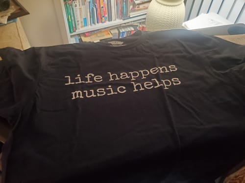 Customer's black Life Happens T-Shirt laid out, showing the white text 'life happens music helps'.
