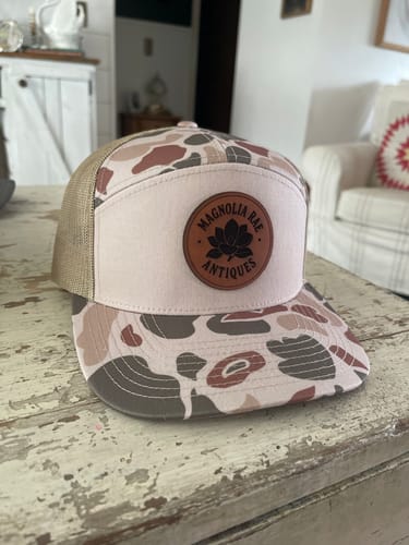 Customer's custom trucker hat with a pink camo pattern, tan mesh, and a round leather patch on a table.