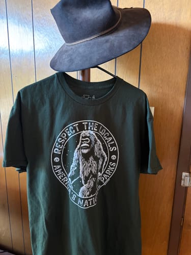 Customer's forest green Respect The Locals Bigfoot Tee with a crisp white graphic, displayed on a hanger.