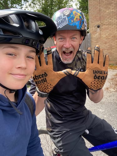 A reviewer holds up the palms of their tan Wraith | Gravity Gloves, showing the text design while outdoors with another person.