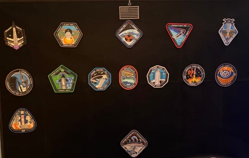 A reviewer's collection of space mission patches, including the Starship Flight 12 patch, on a black board.