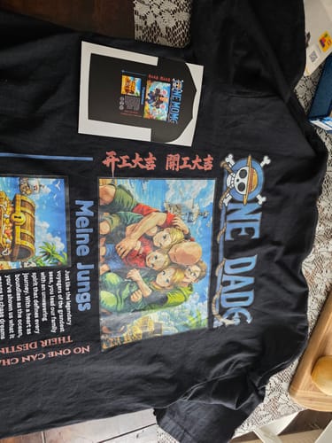 Ein Kunde zeigt das personalisierte schwarze 'ONE DAD'-T-Shirt mit einem farbenfrohen Anime-Aufdruck, der auf einem Tisch liegt.