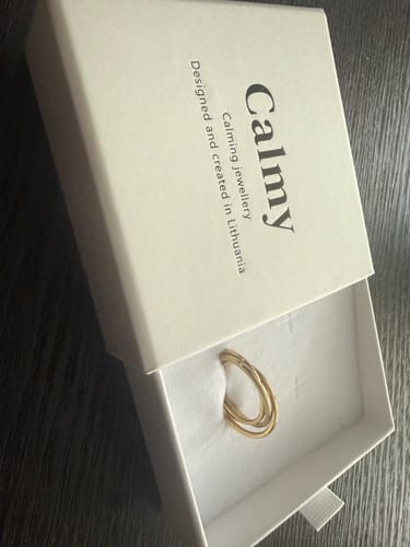 Pirkėjo auksinės spalvos harmonijos žiedas „Calmy“ išpakuojamas iš baltos firminės dėžutės.