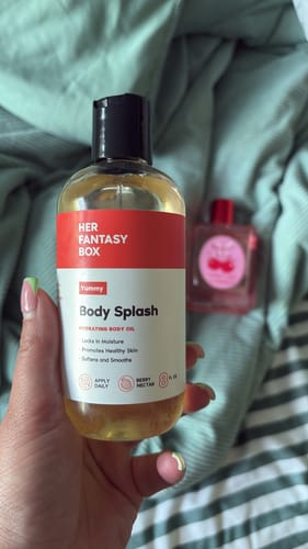 A reviewer's hand holding a clear bottle of the golden Yummy Body Splash oil against a green bedspread.