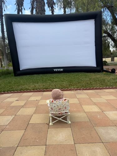 Customer's baby sits in the Clementine portable booster chair on a patio, facing an outdoor movie screen.