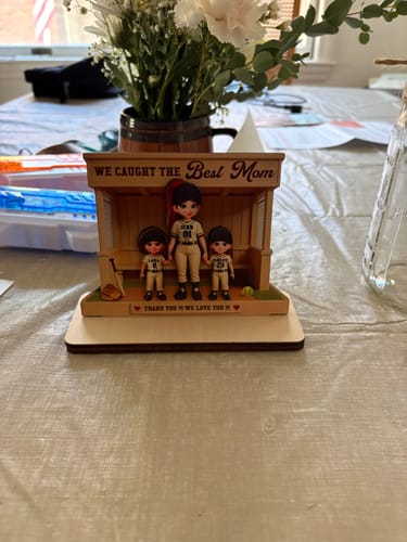 Customer's personalized wooden plaque with a baseball dugout scene featuring a mom and two kids, displayed on a table.
