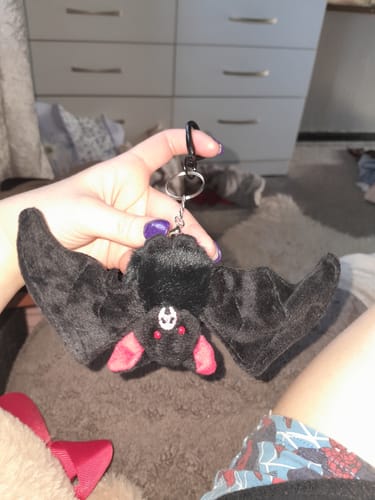 Customer holding the black bat baby keychain. The plush bat has its wings open, showing red eyes and a symbol on its forehead.