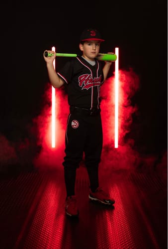 Customer's child posing in the custom black and red baseball jersey while holding a bat in a studio.