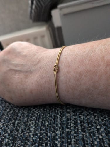 Customer's wrist wearing the delicate gold-colored Forever Knot Bracelet against a dark fabric background.