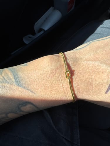 Customer's tattooed arm wearing a delicate gold-colored knot bracelet.