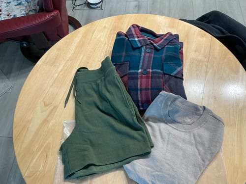 Customer's clothing from a Mystery Bag, including a plaid shirt, green shorts, and a sweater, on a table.
