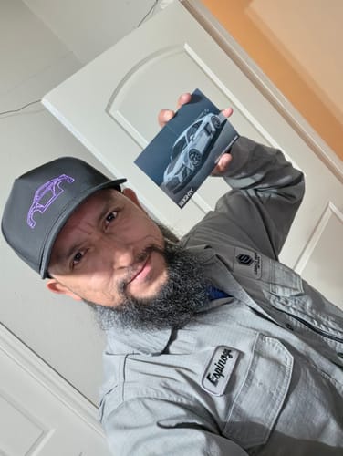 Reviewer in a dark grey DCG88 hat with purple car embroidery, holding up a photo of a sports car.