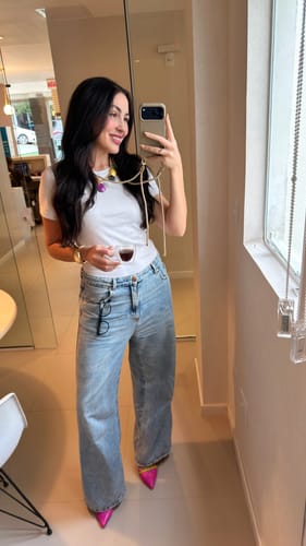 Avaliadora tira uma selfie no espelho, usando sua case Nora Areia DOURADA com a alça fina no pescoço.