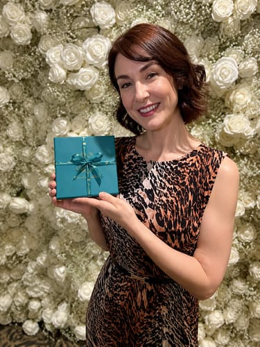 A smiling reviewer holds up the teal box for the Discovery Set in front of a wall of white flowers.